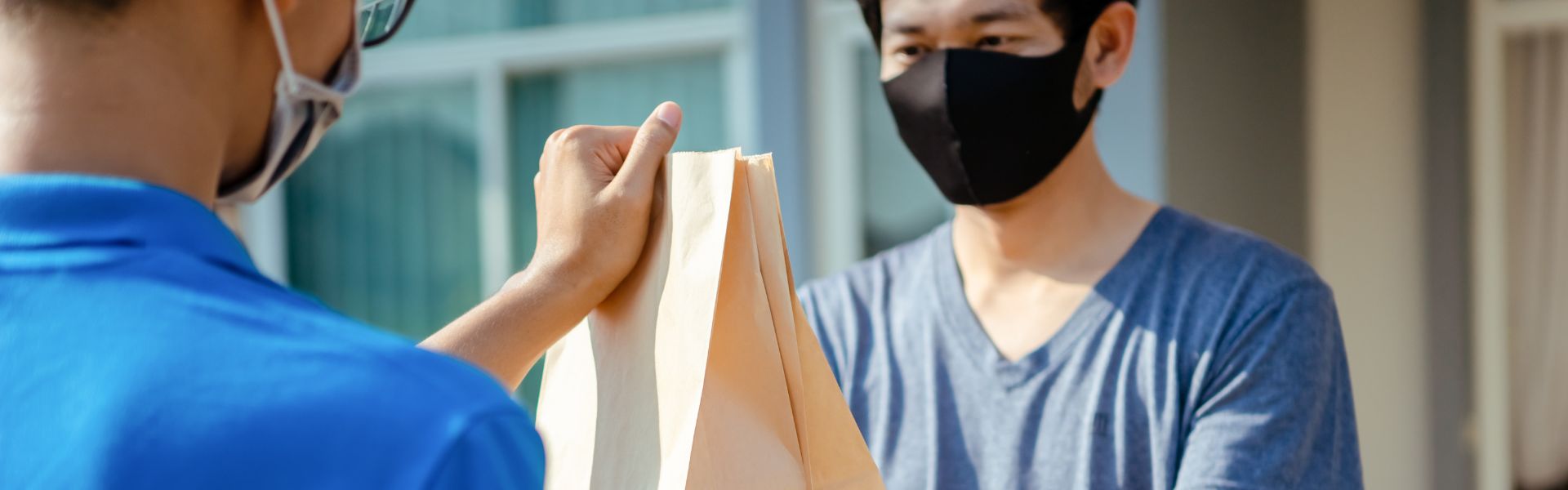 person wearing a blue shirt is handing off a brown paper bag to a person wearing a blue shirt and a black face mask